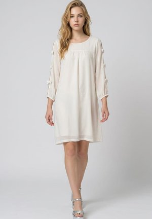 Joven mujer con largo cabello rubio, vestida con un vestido blanco hasta la rodilla con lazos en las mangas y sandalias plateadas de tacón alto, de pie contra un fondo gris.
