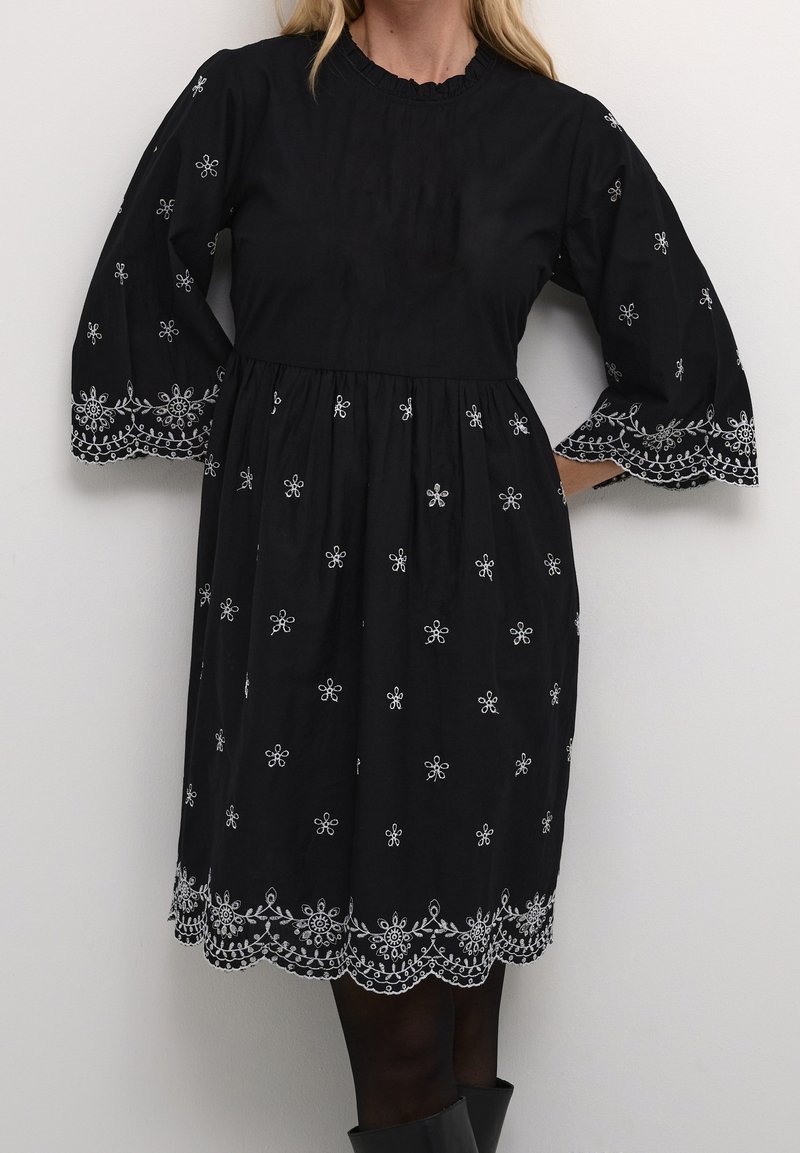 Robe en coton noire avec manches longues bouffantes, ornée de broderies florales blanches et d'un ourlet festonné. Silhouette trapèze, longueur au genou.