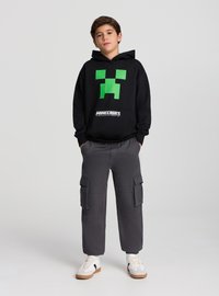 Felpa nera con cappuccio caratterizzata da un design con volto verde pixelato, abbinata a pantaloni cargo grigi e sneaker bianche con accenti neri.
