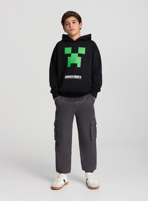 Felpa nera con cappuccio caratterizzata da un design con volto verde pixelato, abbinata a pantaloni cargo grigi e sneaker bianche con accenti neri.