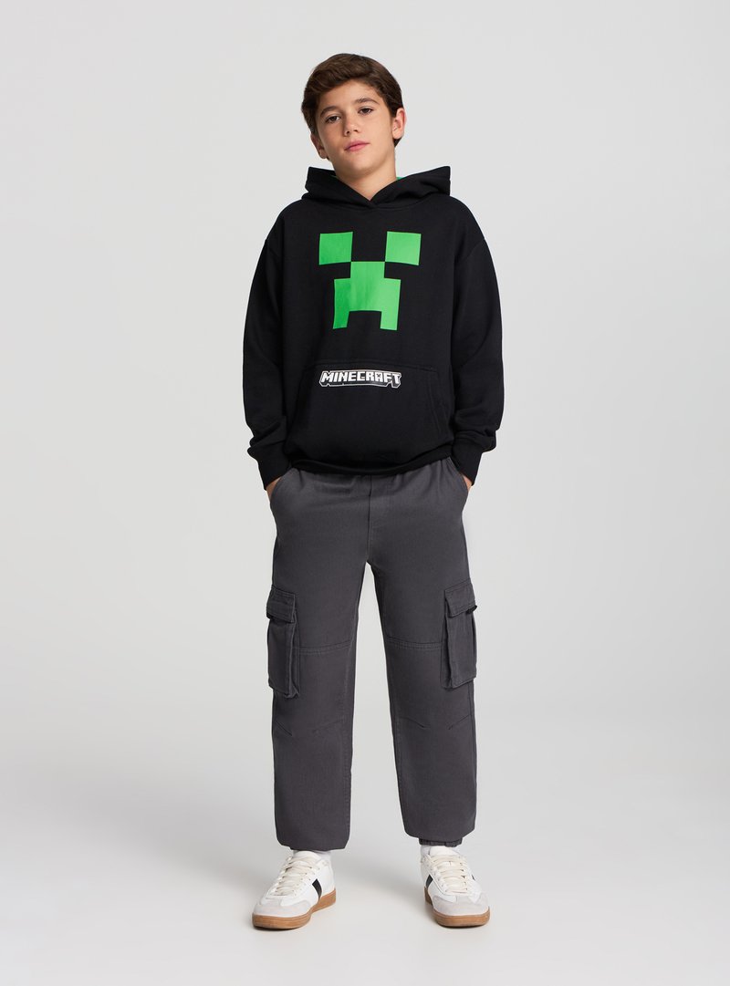 Felpa nera con cappuccio caratterizzata da un design con volto verde pixelato, abbinata a pantaloni cargo grigi e sneaker bianche con accenti neri.