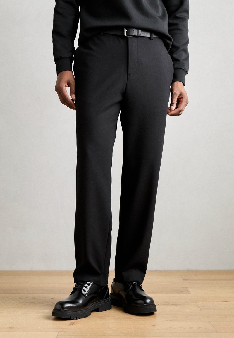 Personne portant un pantalon noir à jambes droites, une ceinture noire, des chaussures habillées noires à lacets et un haut noir à manches longues, debout sur un sol en bois.