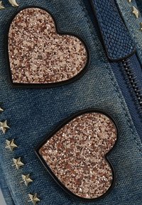 Tela vaquera con dos parches de corazones en brillo rosa dorado, contornos negros y adornos de estrellas doradas en el lado izquierdo. Detalle de cremallera visible.