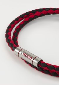 Roter und schwarzer geflochtener Lederarmband mit einem silbernen Metallschluss, auf dem "HUGO" in Rot geprägt ist. Glatte Textur und klare Linien.