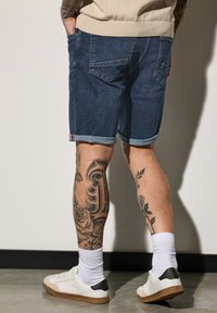 Shorts de mezclilla en azul oscuro con dobladillo enrollado, que cuentan con cuatro bolsillos, llevados con zapatillas blancas y calcetines de tobillo con un diseño.