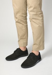 Černé semišové boty s texturovaným povrchem, zdobené hladkým koženým akcentem, šňůrkami a kulatou špičkou. Nosit s béžovými zkrácenými kalhotami.