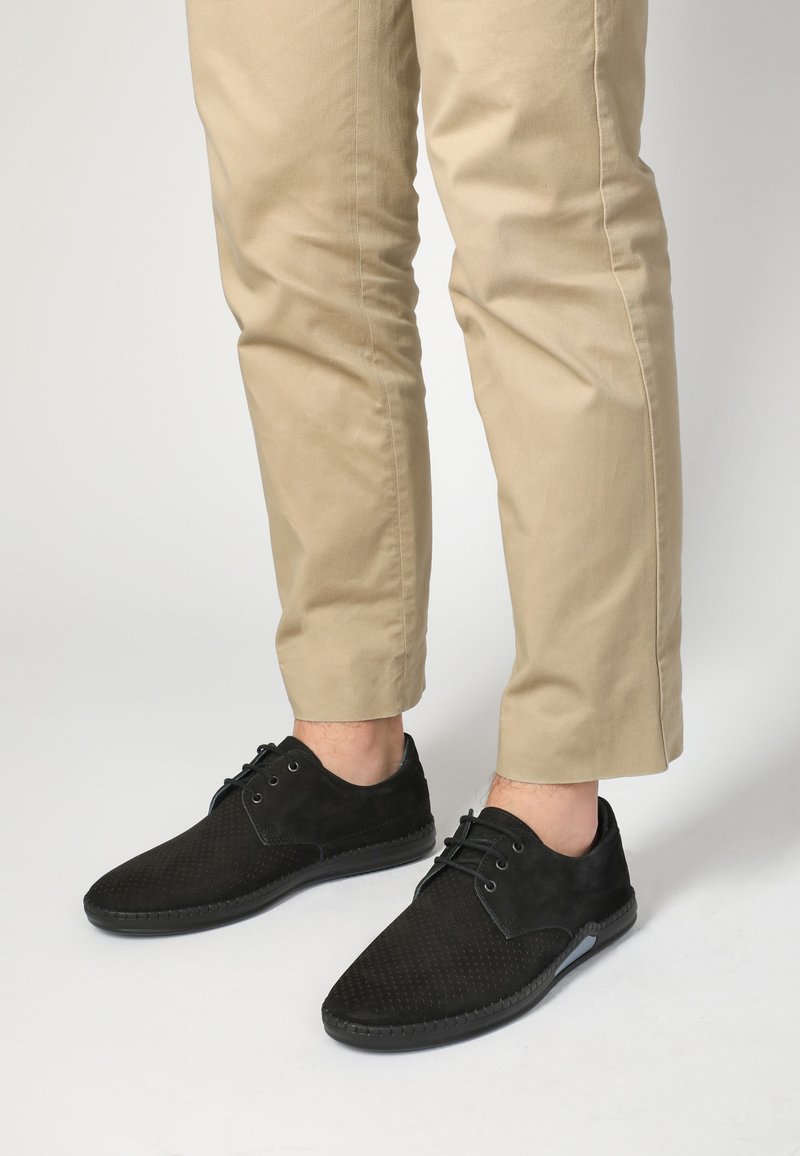 Černé semišové boty s texturovaným povrchem, zdobené hladkým koženým akcentem, šňůrkami a kulatou špičkou. Nosit s béžovými zkrácenými kalhotami.