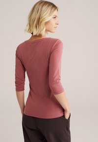 Camisetta rosa chiaro a tre quarti in tessuto morbido con scollatura rotonda. Vista da dietro, abbinata a pantaloni scuri.