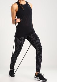 Nainen mustissa treenivaatteissa tekee vastusnauhakoukkua seisten jalat eristyksissä nauhassa.