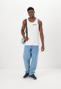 Top din bumbac alb cu logo-ul negru "RIPCURL", asociat cu blugi largi de culoare bleu deschis și adidași negri, ținând o geantă mică de umăr albastră.