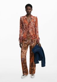 Blusa de manga larga, abierta por delante, y pantalones anchos con un estampado floral en naranja y marrón. La modelo sostiene una chaqueta de mezclilla azul. Un calzado blanco completa el conjunto.