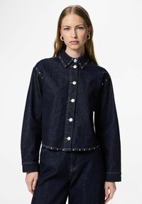 Mørk denimjakke med krage, frontknapper og sølvfargede naglepynt langs skuldrene og kanten. Har lange ermer og kort passform.