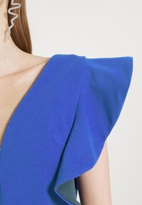 Vestido azul com mangas volumosas e decote em V profundo, feito de um tecido texturizado. Realçado com um brinco espiral.