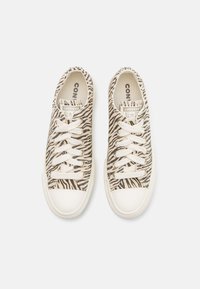 Μπεζ αθλητικά παπούτσια με σχέδιο μαύρης ζέβρας. Επάνω μέρος από καμβά, λευκή καουτσούκ σόλα και λευκοί κορδόνια. Περιλαμβάνει το λογότυπο 'Converse All Star'.