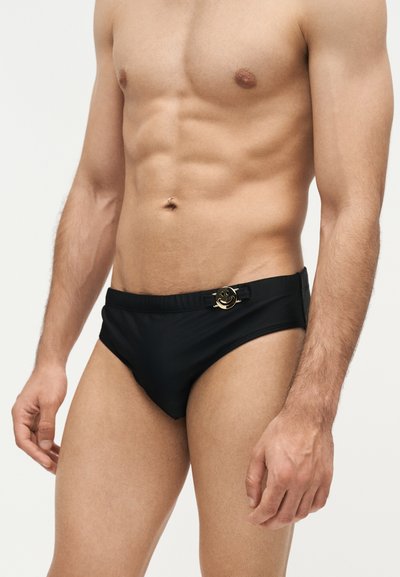 Άνω μέρος ανδρικού σώματος που φοράει μαύρα μαγιό με μεταλλική αγκράφα με ένα χαμογελαστό πρόσωπο στη μέση, στέκεται μπροστά σε ένα απλό φωτεινό φόντο.