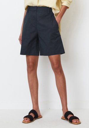Personne portant un short noir au genou, une chemise jaune clair et des sandales noires tissées, debout contre un mur blanc.