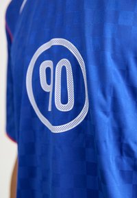 Zils sporta krekls ar šaha spēles rakstu, ar izteiktu baltu apļa emblēmu, kurā ir numurs "90" ar sietveida detaļām. Sarkani akcenti apkaklē.
