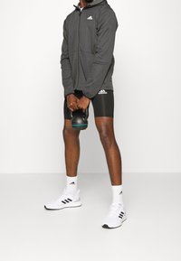 Sudadera con cremallera gris con detalles en negro, pantalones cortos negros, zapatillas blancas y una pesa rusa. Presenta el logo de Adidas en la ropa.