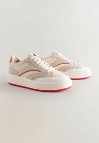 Sneaker beige con suole bianche e dettagli rossi. Presentano materiali in suede e pelle liscia, punta tonda e lacci piatti.