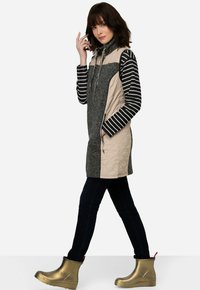 Gequilte vest in grijs en beige met een hoge kraag, gecombineerd met een gestreept overhemd met lange mouwen. Zwarte jeans en gouden regenlaarzen maken de look compleet.