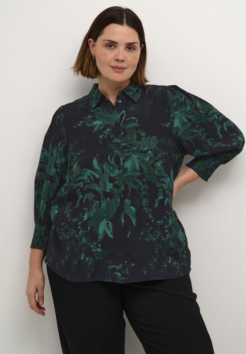 Dunkles Hemd mit grünem Blumenmuster, Knopfleiste, und drei Viertel Puffärmeln mit gerüschten Bündchen. Glatte Stofftextur.