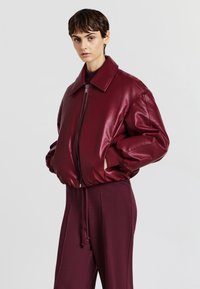 Giubbotto bomber in ecopelle bordeaux con ampio colletto, polsini a costine e chiusura con zip. Indossato sopra pantaloni bordeaux coordinati.