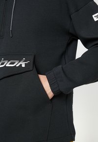 Svart huvtröja med känguruficka, med en vit "Reebok"-logotyp på framsidan och strukturerade ärmar.