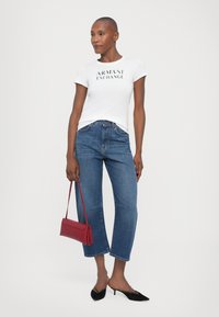 Hvit t-skjorte med "ARMANI EXCHANGE" trykk, blå høyt liv jeans, svarte spisse ballerinasandaler og en rød håndveske. Enkelt, moderne design.