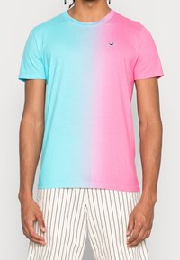 Man med en kortärmad t-shirt i en blå-till-rosa gradient och vita byxor med svarta vertikala ränder, stående mot en neutral bakgrund.