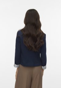 Jūras zilās krāsas žakete ar svītrainām piedurknēm, pielāgota fit, teksturēta auduma, valkāta kopā ar bēšiem augstām vidukļa biksēm. Aizmugures skats, kurā redzami viļņaini mati.