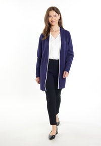 Cardigan lungo viola con rifiniture bianche, indossato sopra una camicetta bianca e pantaloni neri. Il modello indossa tacchi neri e sta camminando in avanti.
