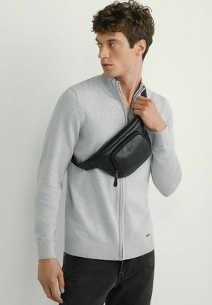 Młody mężczyzna w jasnoszarym swetrze na zamek i czarnej skórzanej torbie na ramię, patrzący na bok na jednolitym tle.