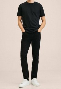 Camiseta de algodón negra con cuello redondo, combinada con unos pantalones vaqueros negros, ambos ajustados. Zapatillas blancas completan el conjunto, de pie contra un fondo neutro.