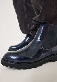 Stivali blu navy lucidi con una superficie liscia, pannelli laterali elasticizzati e suole nere robuste, parzialmente coperti da jeans scuri.