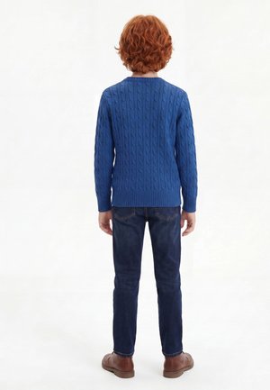 Kind mit lockigem roten Haar, das einen blauen Zopfpullover, dunkle Jeans und braune Schuhe trägt, steht mit dem Rücken zur Kamera vor weißem Hintergrund.