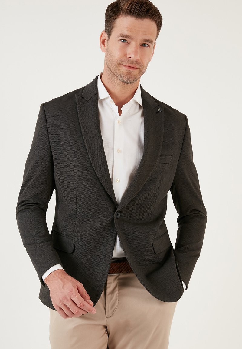 Blazer grigio scuro in tessuto testurizzato, chiusura a bottone singolo, revers a V e due tasche frontali. Indossato sopra una camicia bianca.