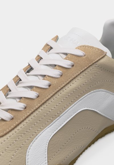 Gros plan d'une chaussure de sport en cuir beige et blanc avec des détails en daim beige et des lacets blancs sur un fond clair.
