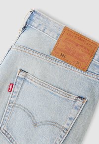 Ανοιχτό μπλε τζιν παντελόνι με ταμπέλα από ταμπά δέρμα που φέρει κείμενο. Κανονικός σχεδιασμός με πέντε τσέπες, αντίθετη ραφή και κόκκινη ταμπέλα.