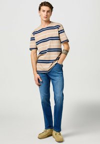 Camiseta de manga corta a rayas en beige y azul marino, jeans azules, zapatos beige. El modelo está de pie con las manos en los bolsillos, mirando hacia adelante. Diseño minimalista.
