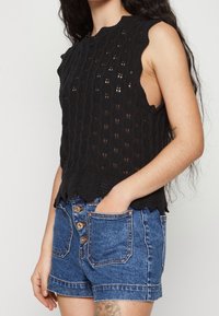 Černý pletený top bez rukávů s texturovaným vzorem, vroubkovanými okraji a otevřenými boky, spárovaný s džínovými šortkami s vysokým pasem a zapínáním na knoflíky.