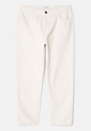 Mies, jolla on beige pitkähiha-Henley-paita, luonnonvalkoiset corduroy-housut ja valkoiset tennarit, seisoo yksinkertaisen vaaleanharmaan seinän edessä.