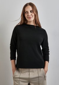 Schwarzes Langarmoberteil mit Rundhalsausschnitt, aus strukturiertem Stoff und mit taillierten Ärmeln, kombiniert mit beigen hochtaillierten Hosen.