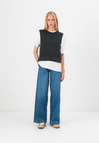 Gilet en tricot gris avec une texture côtelée porté sur un T-shirt blanc, associé à un jean large bleu et des chaussures beiges. Design simple et superposé.