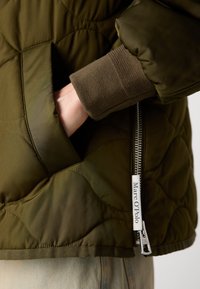 Quiltet olivgrønn jakke med ribbede ermer, sidelomme og sølvglidelås med en merkelapp med "Marc O'Polo"-merke.