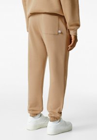 Beige Jogginghose mit schmalen Bündchen und Gesäßtasche. Hergestellt aus weichem Stoff, bietet sie ein minimalistisches Design und ein kleines Etikett an der Tasche.