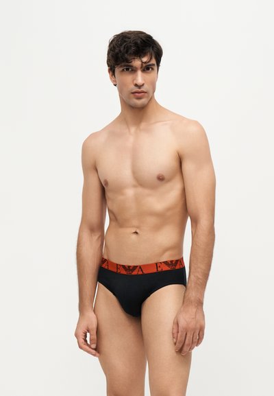Jeune homme aux cheveux courts et foncés portant un slip noir avec une ceinture rouge avec un logo, debout sur un fond clair uni.