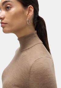 Brązowy sweter o wysokim kołnierzu wykonany z miękkiej dzianiny, o dopasowanym kroju i subtelnej struktury prążkowej. Małe kolczyki w kształcie kółek uzupełniają całość.