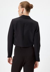Zwarte cropped blazer met lange mouwen, getailleerde pasvorm, subtiele textuur en een enkele naden aan de achterkant. Gecombineerd met zwarte broeken.