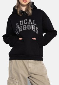 Czarna bluza z kapturem, wykonana z miękkiego materiału, z dużym tekstem ozdobionym dżetami "LOCAL HEROES" na klatce piersiowej. Zawiera kieszeń kangurkę.