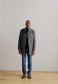 Cappotto grigio in lana con collo alto, chiusura a bottoni e due grandi bottoni. Camicia nera, jeans blu e scarpe grigie. In piedi su un pavimento di legno.